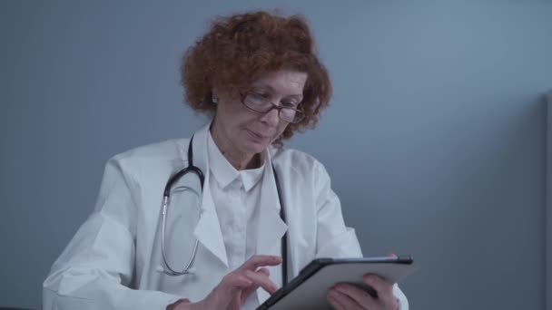 Пожилая женщина профессиональный врач с использованием цифрового планшета в больнице. Врач держит современное технологическое устройство, работающее в режиме онлайн с помощью приложений дистанционной телемедицины. Концепция технологии здравоохранения - Кадры, видео
