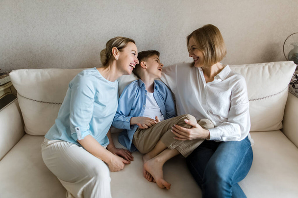 Two adult women and a child are sitting on the couch, chatting and smiling. - Zdjęcie, obraz