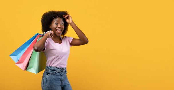 Seizoensverkoopconcept. Verrast zwarte vrouw met kleurrijke boodschappentassen, gele achtergrond, panorama, vrije ruimte - Foto, afbeelding