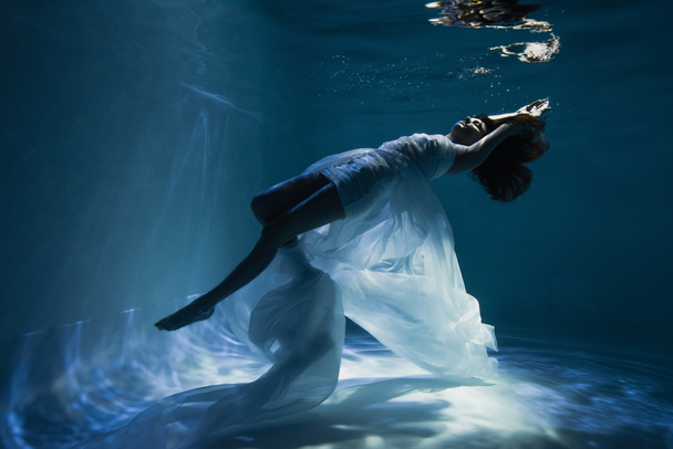 lighting on graceful young woman in white elegant dress swimming in pool  - Foto, Imagem