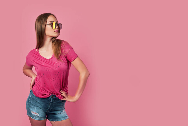 Photo shot of pretty young woman isolated over pink background wall wearing casual pink t-shirt and stylish colourful sunglasses looking at camera - Foto, afbeelding
