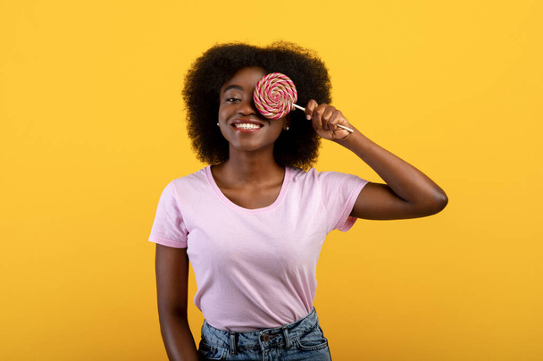 Sladký zub. Pozitivní mladá africká americká žena pokrývající jedno oko s lízátkem přes jasně žluté studio pozadí - Fotografie, Obrázek