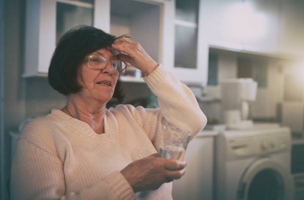Mujer mayor con dolor de cabeza y píldora para beber en la cocina - Foto, imagen