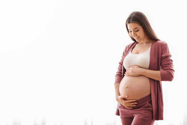 Young woman expecting baby embracing belly while standing near window at home - Foto, Bild