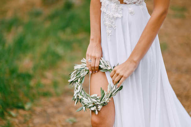 Schöne Braut in einem weißen bestickten Kleid hält einen Kranz aus Olivenblättern. Das Bein der Braut ist durch den Schnitt des Kleides sichtbar - Foto, Bild