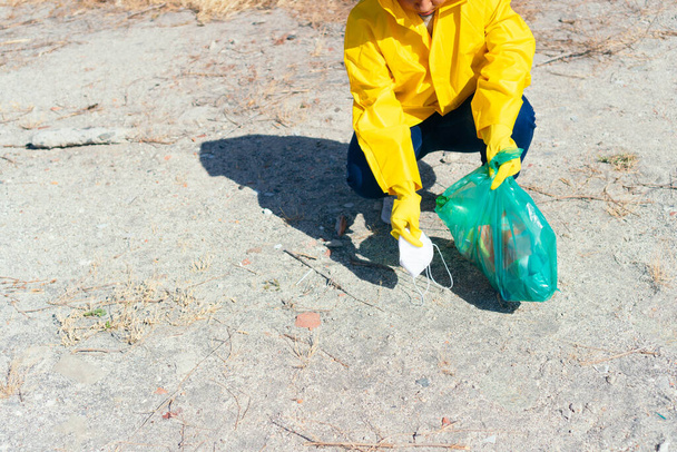 Woman collecting disposable face mask in garbage bag while cleaning beach - Fotoğraf, Görsel