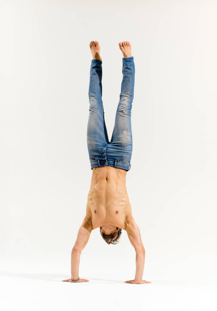 Athletic man in jeans with bare chest performing handstand on a white background. - Photo, image