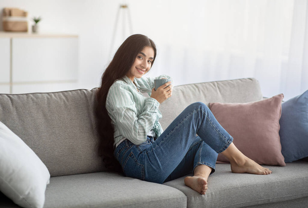Full length portrait of millennial Indian lady drinking coffee, resting on sofa, smiling at camera, indoors - Photo, Image