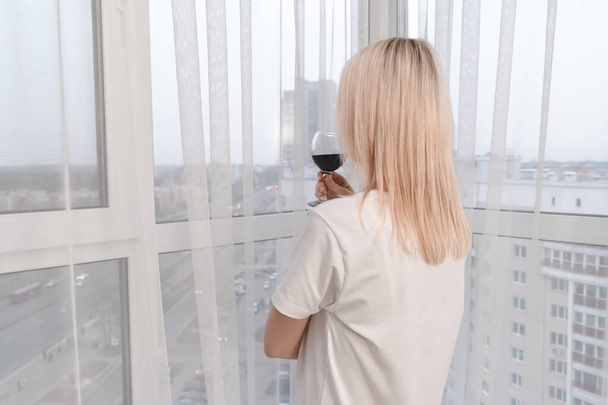 Mulher loira jovem em uma camiseta branca de tamanho grande com um copo de vinho tinto fica na janela e olha para a distância da cidade. Grandes janelas no quarto. Foto de trás - Foto, Imagem