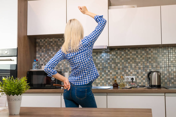 Cheerful young slender blonde woman in blue jeans and a shirt cooks in the kitchen and dances, copy space. Photo from the back - Fotó, kép