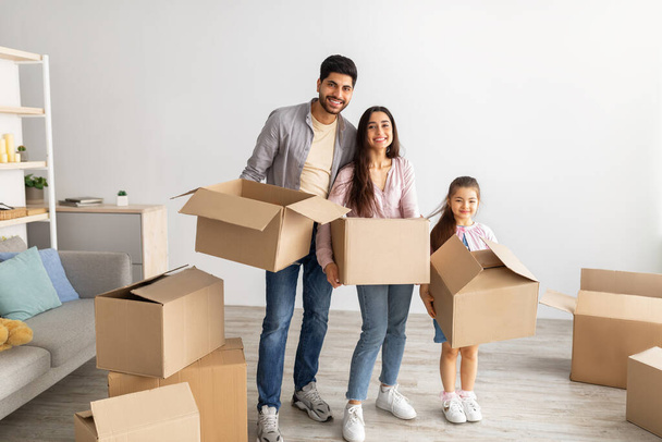 Happy eastern family holding carton boxes and smiling at camera, standing in their new apartment - Foto, Bild