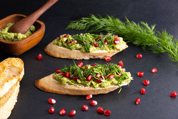 avocado snack, sprinkled with ripe pomegranate seeds, sesame seeds and fragrant fresh dill, black background - Photo, Image