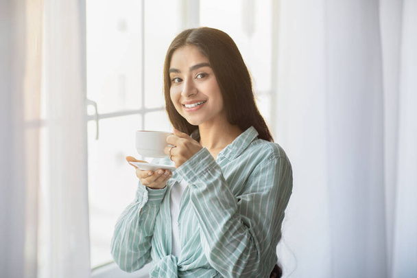 Lovely young Indian woman having coffee break in front of window at home - 写真・画像