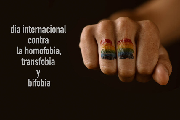 die in die Faust eines jungen Menschen gemalte Regenbogenfahne und der Text Internationaler Tag gegen Homophobie, Transphobie und Biphobie auf Spanisch vor schwarzem Hintergrund - Foto, Bild
