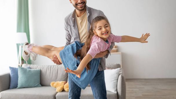 Сімейні ігри. Щасливий молодий батько, що носить маленьку дочку, як літак, тримає дівчину на руках і грає вдома
 - Фото, зображення