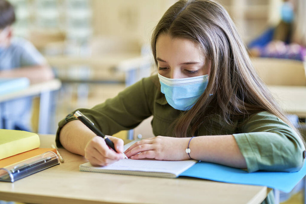 Κοντινό πλάνο της μαθήτριας με προστατευτική μάσκα γραφής στο σημειωματάριο - Φωτογραφία, εικόνα