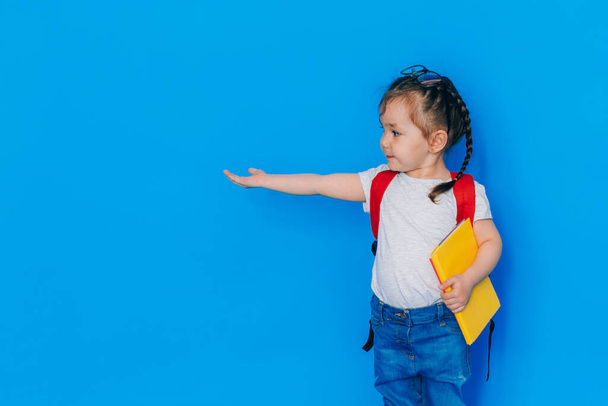 Torniamo al concetto di scuola. Ragazza della scuola con zaino rosso e tenendo cartella gialla davanti a sfondo blu. Il bambino stese la mano destra di lato. - Foto, immagini