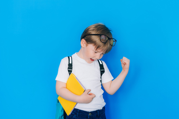 Back to school concept. Schoolboy boy with backpack and holding yellow notebook against blue wall. Child clenched fist showing success. - Fotoğraf, Görsel