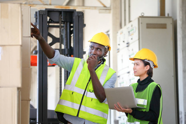 Αφροαμερικανοί και ασιάτες εργαζόμενοι που φορούν γιλέκο ασφαλείας ενώ εργάζονται σε αποθήκη και χρησιμοποιούν walkie talkie έλεγχο για την απογραφή του προϊόντος χρησιμοποιώντας φορητό υπολογιστή - Φωτογραφία, εικόνα