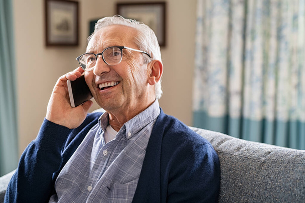 Smiling old man talking over smartphone while relaxing at home. Senior man after retirement using smart phone to connect with friends and family. Carefree elder talking over mobile phone with her daughter while sitting on couch and looking up. - Zdjęcie, obraz