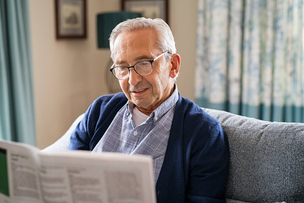 Senior man wearing spectacles reading a newspaper while relaxing at home. Retired man sitting on couch reading newspaper for daily updates. Old man in glasses relaxing at home. - Foto, imagen