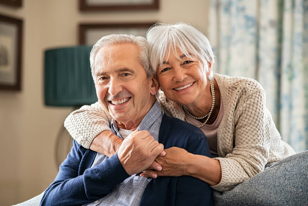 Portrait of romantic senior man with his beautiful wife stay at home. Smiling and caring old woman embracing from behind her retired husband sitting on couch. Cheerful old couple looking at camera with joy. - Foto, imagen