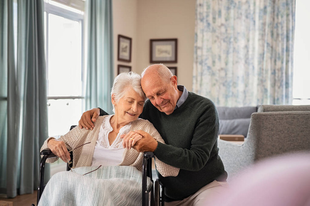 Romantic senior man embracing woman sitting on wheelchair at nursing home. Elderly couple in love embracing with eyes closed and deep respect for each other. Lovely old man holding hands of his disabled wife at care centre during a visit. - Foto, imagen