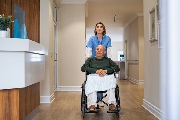 Szczęśliwa pielęgniarka pomaga staruszkowi na wózku inwalidzkim w domu opieki. Młody pracownik służby zdrowia opiekujący się niepełnosprawnym pacjentem w podeszłym wieku w ośrodku opieki. Przyjazny opiekun popycha starego pacjenta siedzącego na wózku inwalidzkim w centrum opieki. - Zdjęcie, obraz