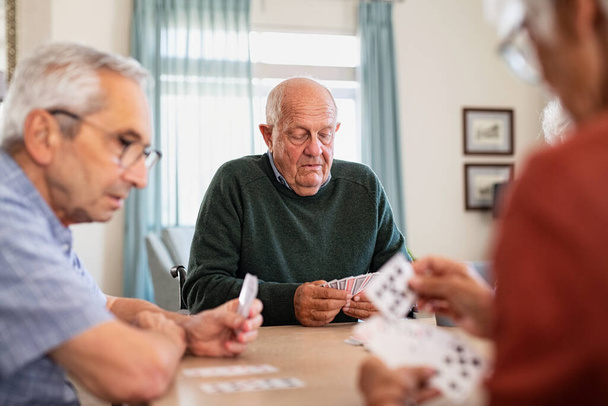 Retired senior man in wheelchair playing cards with his friends at nursing home. Group of old friends playing cards at care centre. Focused senior people playing cards game at lunch table during afternoon. - Fotografie, Obrázek
