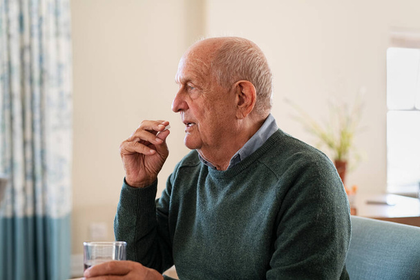 Ill senior man taking medicine at care centre. Elderly man taking pills for depression sitting on couch. Old upset patient swallowing pill with glass of water. - Fotoğraf, Görsel