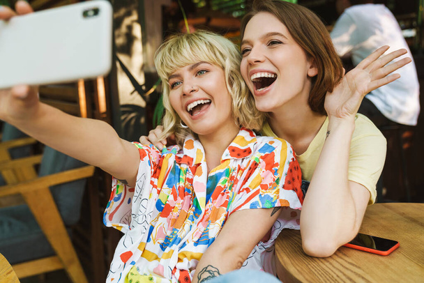 Image of excited two women waving hand and taking selfie photo on smartphone while sitting in cozy cafe outdoors - Valokuva, kuva