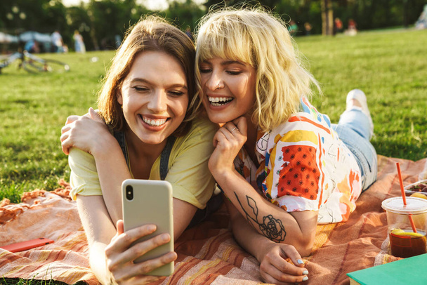 Image of joyful beautiful two women taking selfie on cellphone while have picnic on grass in summer park - Photo, Image