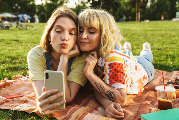 Image of joyful beautiful two women taking selfie on cellphone while have picnic on grass in summer park - Фото, изображение