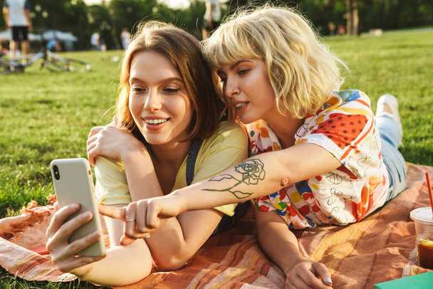 Image of pleased nice two women using and pointing finger at cellphone while have picnic on grass in summer park - Foto, Imagem