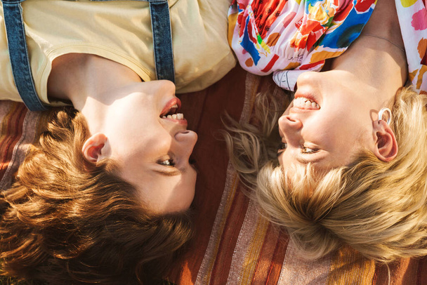 Image of happy two women smiling at each other and lying on blanket while have picnic in summer park - Foto, afbeelding