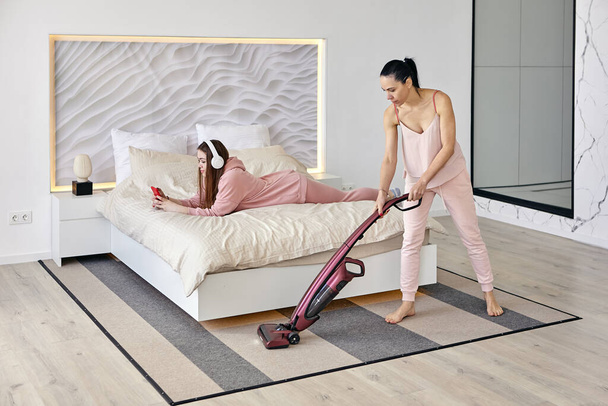 Young woman lies on her stomach and listens to music while her mother uses cordless vacuum cleaner to dust floor. - Photo, image