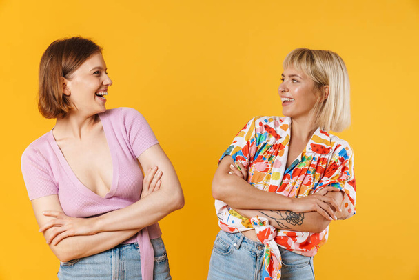 Portrait of two cheerful lovely girlfriends wearing summer clothes standing isolated over yellow background, arms folded - Φωτογραφία, εικόνα