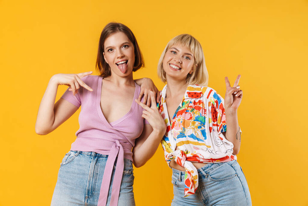 Portrait of two cheerful lovely girlfriends wearing summer clothes standing isolated over yellow background - Fotó, kép