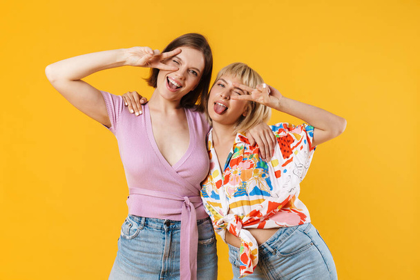 Portrait of two cheerful lovely girlfriends wearing summer clothes standing isolated over yellow background, showing peace geasture - Valokuva, kuva