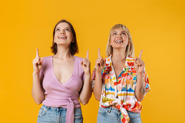 Portrait of two cheerful lovely girlfriends wearing summer clothes standing isolated over yellow background, pointing fingers up - 写真・画像