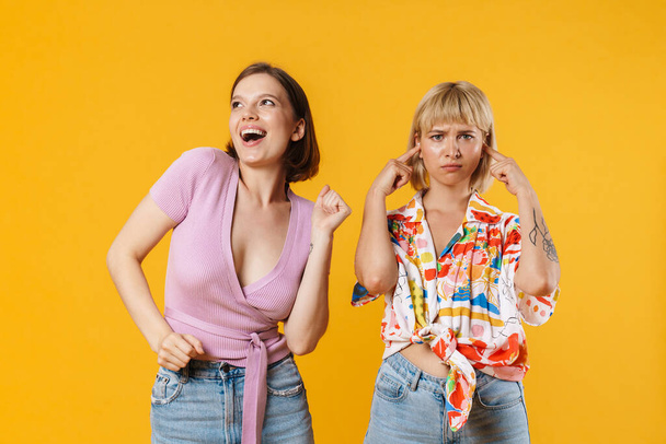 Portrait of two cheerful lovely girlfriends wearing summer clothes standing isolated over yellow background, having fun, dancing - Photo, image