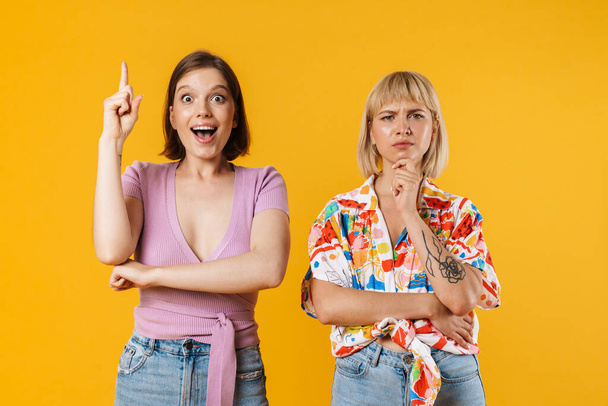 Portrait of two cheerful lovely girlfriends wearing summer clothes standing isolated over yellow background, having idea - Фото, изображение