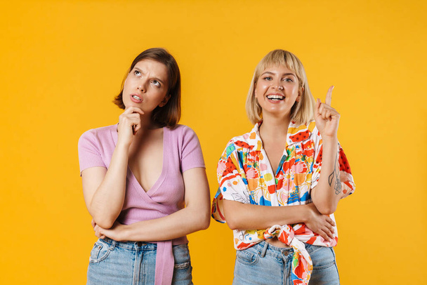 Portrait of two cheerful pensive lovely girlfriends wearing summer clothes standing isolated over yellow background, having idea - Fotografie, Obrázek