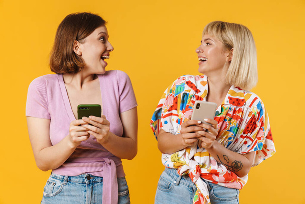 Portrait of two cheerful lovely girlfriends wearing summer clothes standing isolated over yellow background, holding mobile phone - Foto, imagen