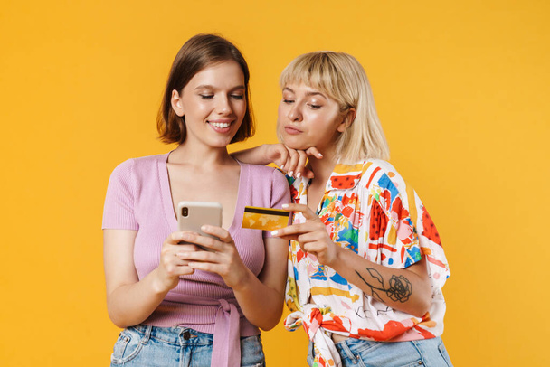 Portrait of two cheerful lovely girlfriends wearing summer clothes standing isolated over yellow background, holding mobile phone, shopping online with credit card - 写真・画像