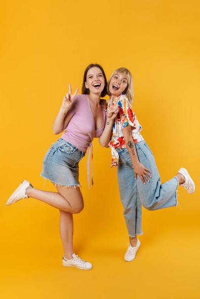 Full length portrait of two cheerful lovely girlfriends wearing summer clothes standing isolated over yellow background, having fun - Fotoğraf, Görsel