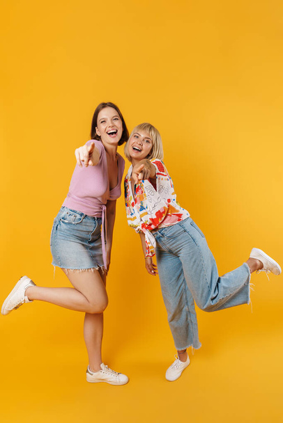 Full length portrait of two cheerful lovely girlfriends wearing summer clothes standing isolated over yellow background - Photo, Image