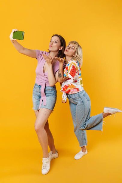 Full length portrait of two cheerful lovely girlfriends wearing summer clothes standing isolated over yellow background, taking a selfie - Foto, afbeelding