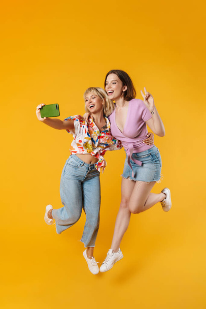 Full length portrait of two cheerful lovely girlfriends wearing summer clothes jumping isolated over yellow background, taking a selfie - Zdjęcie, obraz