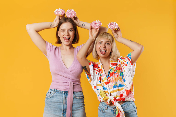 Portrait of two cheerful lovely girlfriends wearing summer clothes standing isolated over yellow background, showing tasty donuts, having fun - Foto, Imagen
